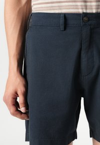Şorturi bleumarin dintr-un material texturat, cu închidere prin buton, buzunare laterale și cusături vizibile pe margine și talie.