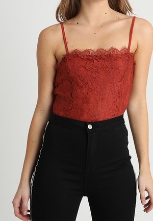 Frau trägt ein ärmelloses, rostrot-spitzenbesetztes Camisole mit dünnen Trägern und hoch taillierte schwarze Hose mit weißen Seitenstreifen.