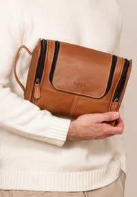 Tan leather bag with black zippers, featuring a flap closure and external stitching details. Compact, rectangular design with a handle.