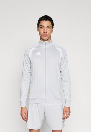 Chaqueta deportiva gris claro con cremallera, cuello alto, paneles blancos a contraste y logo negro de Adidas en el pecho izquierdo, combinada con pantalones cortos a juego.