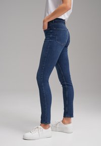 Personne portant un jean skinny bleu foncé et des baskets blanches, debout de côté avec les mains dans les poches devant un fond gris uni.