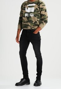 Felpa camouflage nei toni del verde e marrone con grafica bianca "RAW"; jeans skinny neri; scarpe nere con lacci; look casual.
