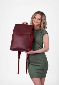Bordová kožená batoh s přehnutým víkem, látkovými bočními kapsami, nastavitelného popruhy a minimalistickým logem na přední straně.