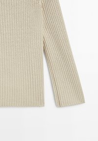 Lang beige strikket ærme med vertikalt ribmønster på hvid baggrund.