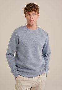 Un hanorac din bumbac de culoare bleu deschis, cu guler rotund, manșete și tiv cu nervuri, având o textură subtilă și o croială lejeră. Asortat cu pantaloni bej.