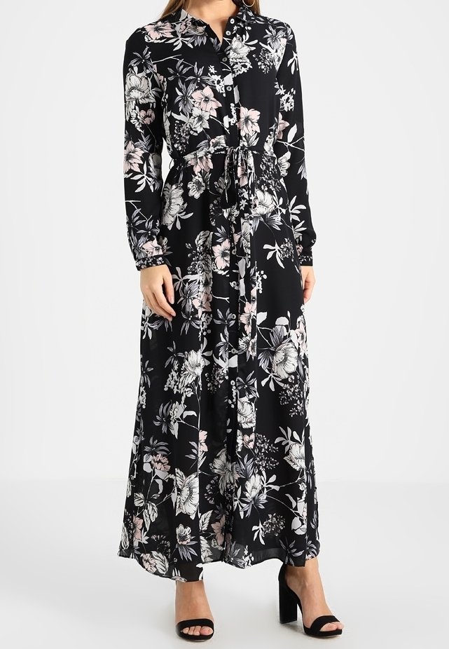 Robe longue à manches longues en tissu noir, ornée de motifs floraux roses et blancs, boutonnée à l'avant, avec un ceinturage à nouer à la taille. Talons hauts visibles.