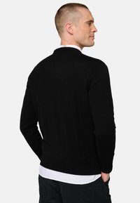 Schwarzer Strickpullover mit Rundhalsausschnitt, langen Ärmeln und weicher Textur, über einem weißen Hemd getragen. Einfaches Design, ohne sichtbare Muster.