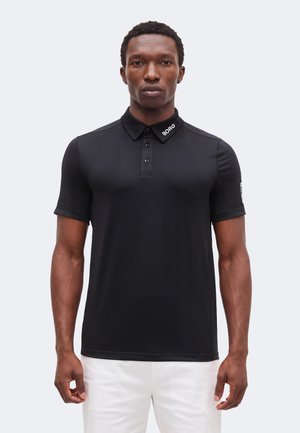 Άνδρας που φοράει εφαρμοστό μαύρο πόλο με το κείμενο «BORG» στον γιακά και στο αριστερό μανίκι, φορώντας άσπρο παντελόνι, στέκεται μπροστά σε απλό φόντο.