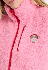 Różowa, miękka kurtka polarowa z wysokim kołnierzem, czerwonym suwakiem oraz naszywką z logo przedstawiającym górski wzór. Miękka tekstura i swobodny krój.