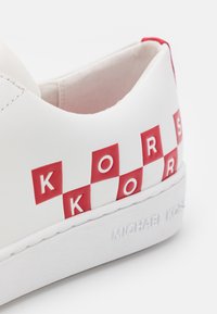 Baskets blanches avec logo rouge et motif à carreaux, fabriquées en matériau synthétique lisse. Dotées d'une épaisse semelle blanche avec le nom de la marque en relief.