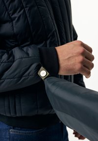 Mörk pufferjacka med quiltat mönster, utrustad med svarta ribbade muddar och ett knäppt band i olivgrönt med silverdetaljer.