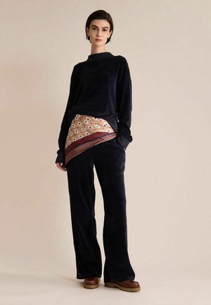 Femme portant un haut noir à manches longues et un pantalon avec une écharpe beige et rouge à motifs nouée à la taille, debout devant un fond beige uni.