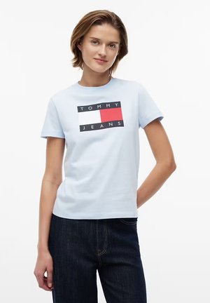 Giovane donna che indossa una maglietta Tommy Jeans azzurra chiara con logo e jeans blu scuro, in piedi con una mano sui fianchi su sfondo bianco.