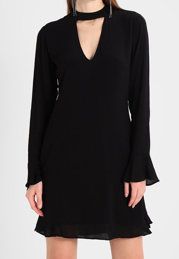 Femme portant une robe noire longue manches avec une découpe en V et des poignets évasés, visible des épaules jusqu'à mi-cuisse.