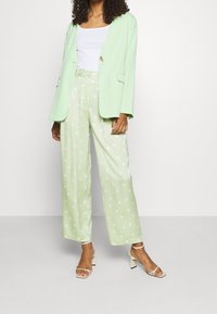 Jasnozielona marynarka z pojedynczym złotym guzikiem, w zestawie z szerokimi spodniami w groszki w jaśniejszym odcieniu zieleni oraz beżowymi sandałami na obcasie.