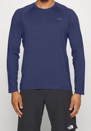 Hombre vistiendo una camiseta de manga larga azul marino y cuello redondo y pantalones cortos negros, ambos con el logo de The North Face, de pie con los brazos relajados a los lados.