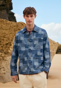 Giovane uomo in piedi su una spiaggia sabbiosa, indossa una giacca di jeans blu a toppe e pantaloni beige, con formazioni rocciose e cielo sullo sfondo.