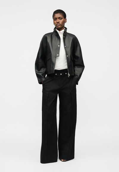 Γυναίκα όρθια με μαύρο δερμάτινο μπουφάν, λευκό ζιβάγκο, μαύρο παντελόνι με φαρδιά μπάρα και ζώνη, τα χέρια στις τσέπες, ουδέτερο φόντο.