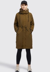 Olivově zelený dlouhý parka s tkaničkami v pase, zapínáním na zip a knoflíky a bočními kapsami, v kombinaci s černými legínami a teniskami.
