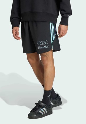 Persoon die zwarte shorts draagt met Revolut- en Audi-logo's, zwarte Adidas-sokken en zwarte Adidas-sneakers met reflecterende strepen.