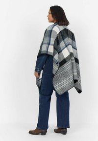 Randig poncho i nyanser av svart, grått och blått. Tillverkad i ett mjukt material med fransade kanter. Bärs över en denimoutfit och ankelskor.