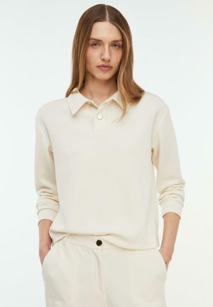 Jeune femme aux cheveux longs et lisses portant un polo à manches longues de couleur crème et un pantalon assorti, mains dans les poches, fond neutre.