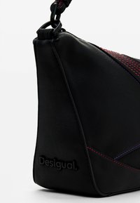 Bolso de cuero negro con forma triangular, con costuras en contraste de color rojo y púrpura y un logo en relieve en el lateral.