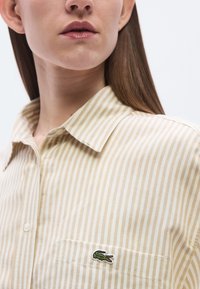Πουκάμισο από βαμβάκι σε μπεζ και κρεμ ρίγες με γιακά, που διαθέτει τσέπη στο στήθος και κεντημένο πράσινο λογότυπο κροκόδειλου στην τσέπη.