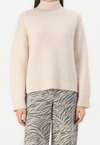 Femme portant un pull à col roulé beige clair et un pantalon gris à motif rayures zèbre noires, debout devant un fond blanc.