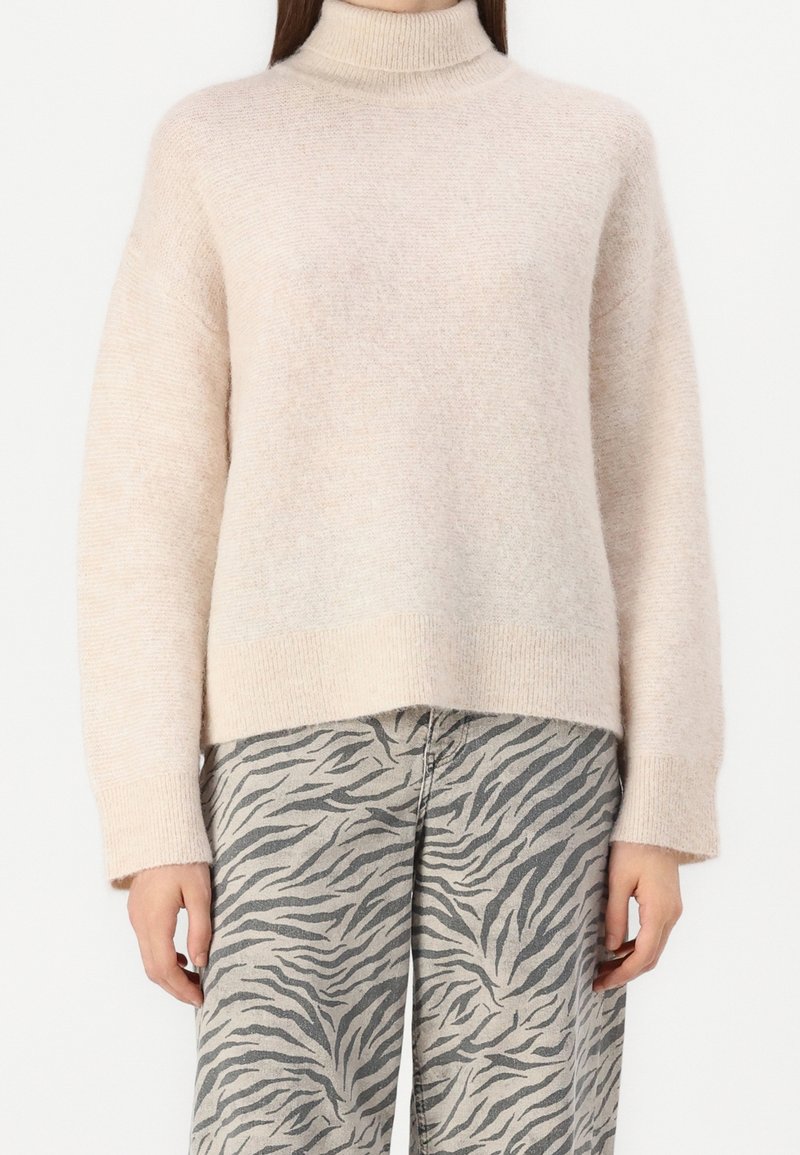 Femme portant un pull à col roulé beige clair et un pantalon gris à motif rayures zèbre noires, debout devant un fond blanc.