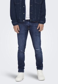Mörkblå denimjacka och jeans med en något sliten textur, tillsammans med vita sneakers, som har knäppningar och fickor.