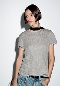 T-shirt grigia a maniche corte in un tessuto morbido e testurizzato, abbinata a jeans di denim azzurro chiaro e una cintura nera. Accentuata da un foulard nero al collo.