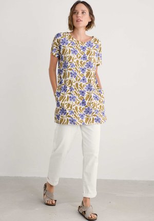 Blumen-Tunika in Beige mit blauen und braunen Blattmustern, kurzen Ärmeln, abgerundetem Saum und zwei Seitentaschen, kombiniert mit weißen Hosen und grauen Sandalen.