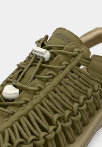 Sandalia verde oliva con cordones elásticos trenzados, cierres de botón blanco y suela acolchada, diseñada para un ajuste ajustable y transpirabilidad.