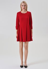 Rode jurk tot op de knie met een ronde hals en driekwartmouwen. Heeft plooien en een lichte structuur. Draagt zwarte ballerina's.