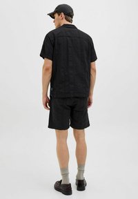 Svart kortärmad skjorta och shorts, strukturerad tyg, avslappnad passform, i kombination med grå strumpor och mörka skor. Modell vänd bort från kameran.