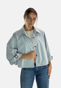 Lysegrå, kort jakke i glat, skinnende stof med stor krave, knaplukning og ærmer med rulleophæng. Båret over en lyse top.