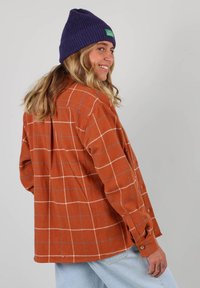 Chemise en flanelle à carreaux orange brûlé, à manches longues, avec un détail de boutons aux poignets et une coupe décontractée, associée à un bonnet en maille violet.