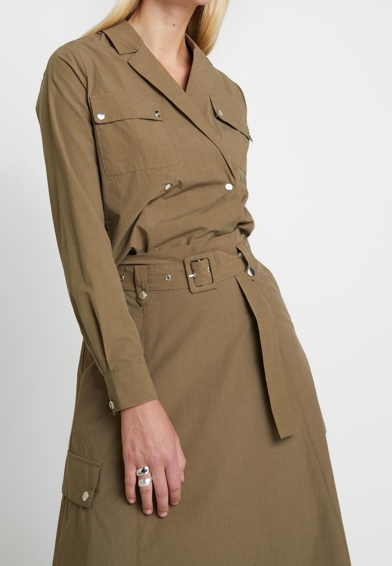 Abito utility khaki con maniche lunghe, vita cintata, tasche sul petto con bottoni e dettagli in argento. Tessuto morbido con vestibilità su misura.