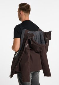 Brun og grå lagdelt parka-jakke med lynlås, hætte og tekstureret stof, holdt over skulderen af en person i en sort skjorte.