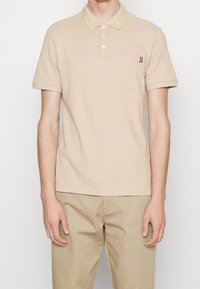 Beige polo tričko vyrobené z bavlny, s knoflíkovou légou, krátkými rukávy a přední kapsou se pruhovaným akcentem.