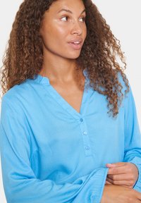 Camisa azul de botones con cuello en V, de manga larga y un acabado texturizado. La tela es ligera, con sutiles patrones de diamantes.