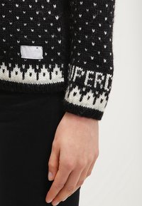 Pletený svetr v čern�é barvě s bílými vzory, s výrazným bílým pruhem a textem "SUPERDRY" na manžetě; měkká, texturovaná tkanina.