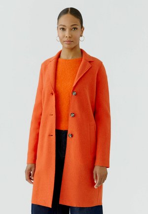 Frau trägt einen leuchtend orangefarbenen, geknöpften Mantel über einem orangenen Pullover und dunklen Hosen, stehend vor einem schlichten hellen Hintergrund.