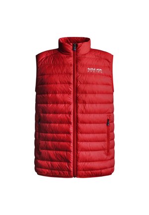 Uomo che indossa un gilet imbottito rosso sopra una maglia da corsa blu navy, pantaloni neri e scarpe da ginnastica bianche, in piedi con una mano in tasca.