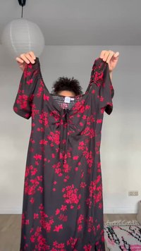 Osoba drži crnu haljinu s crvenim cvjetnim uzorkom, kratkih rukava i detaljem za vezanje u predjelu prsa, u svijetloj sobi s lampionom od papira.