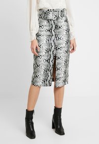 Kvinna som bär en högmidjad, bälteförsedd ormskinnspennkjol med en framklippning, vit blus och svarta klackade ankelboots.