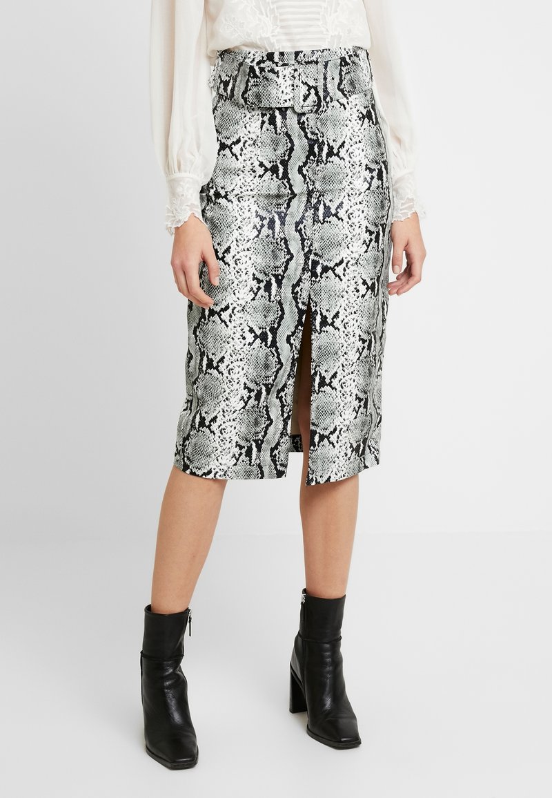 Kvinna som bär en högmidjad, bälteförsedd ormskinnspennkjol med en framklippning, vit blus och svarta klackade ankelboots.