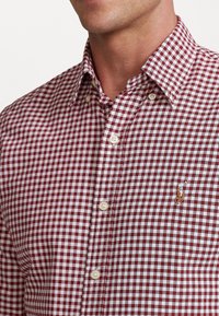Camicia a quadri rossa e bianca con colletto, caratterizzata da un piccolo logo ricamato sul lato sinistro del petto e bottoni bianchi.
