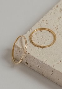 Due anelli d'oro su una superficie di pietra chiara e testurizzata, uno con una fila di piccoli diamanti trasparenti e l'altro una fascia liscia e semplice.